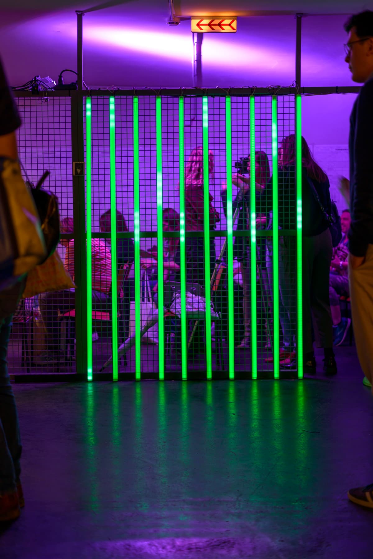 Green Light Tubes on a fence with people behind it during an event